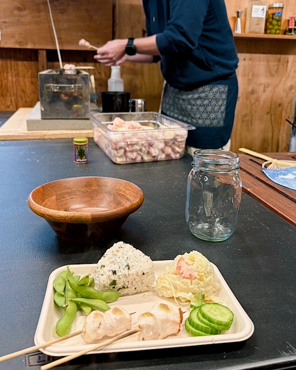 A plate with a rice ball, edamame, pickles, potato salad and two chicken skewers