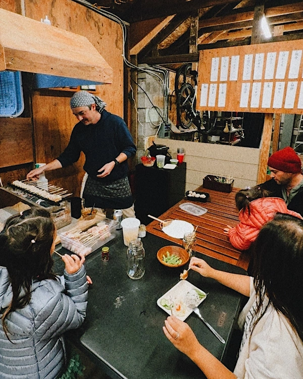 A group of people eating food gathered around a person grill chicken skewers