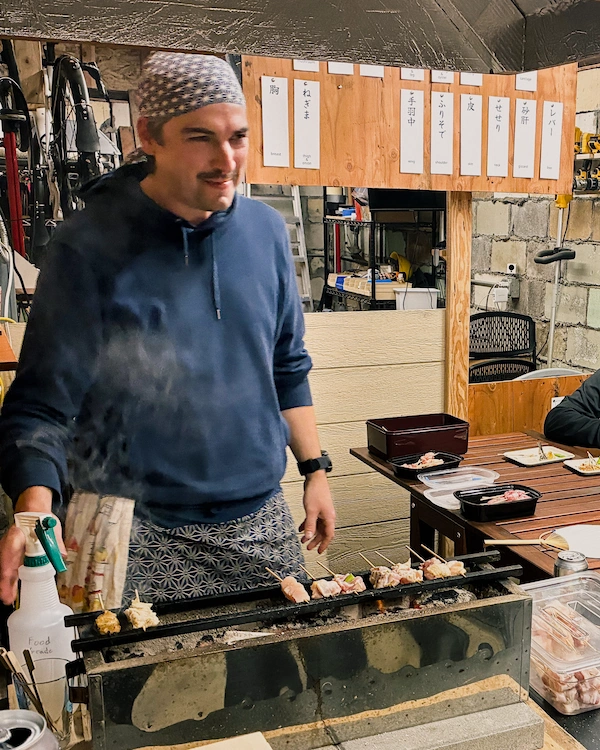A guy smiling while standing in front of some chicken skewers on a japanese style charcoal grill