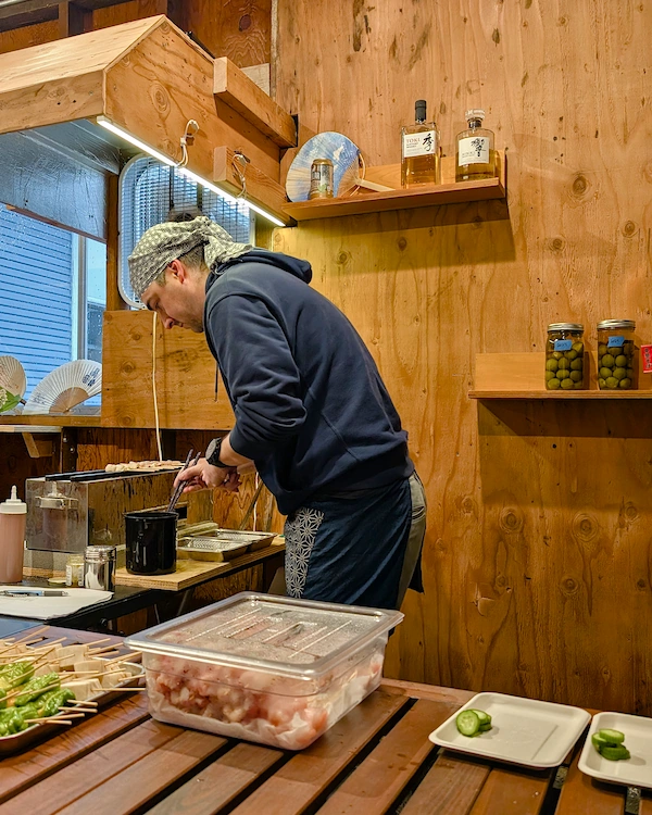 Person grilling skewers in front of a wall will whiskey bottles on it