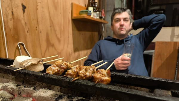 A man relaxes in the background while skewers cook on the grill in the foreground