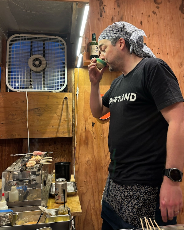 A man standing beside a yakitori grill drinks some sake out of a cup