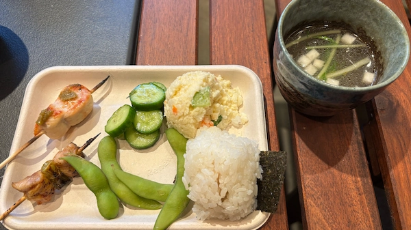Breast and negima skewers along with onigiri, edamame, pickles and potato salad on a plate and a bowl of soup beside the plate