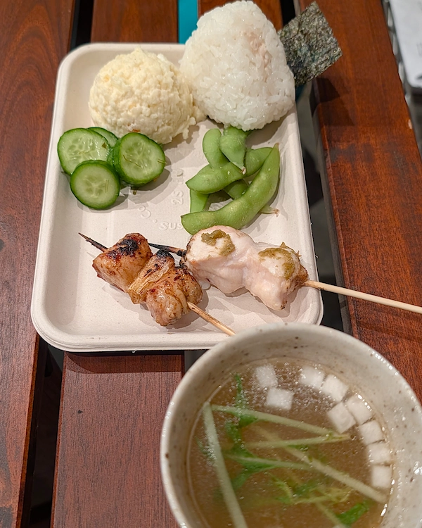 Breast and negima skewers along with onigiri, edamame, pickles and potato salad on a plate and a bowl of soup beside the plate